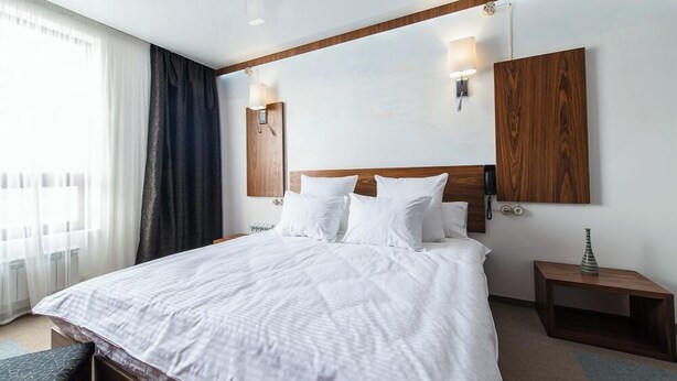 Quarto de hotel com uma cama grande, duas mesas de cabeceira, um painel de madeira na cabeceira e uma janela com cortinas finas.
