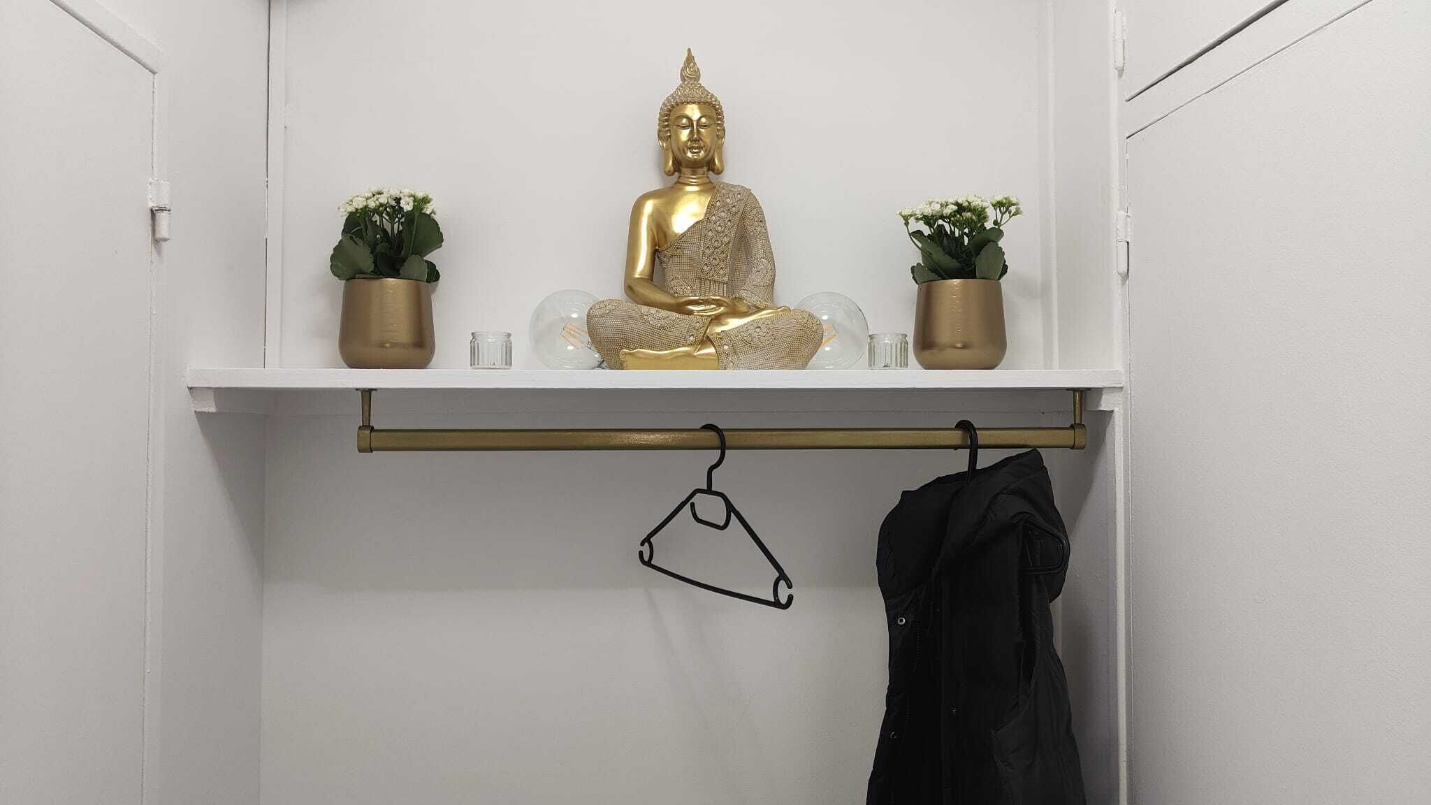 A white room with a shelf holding a golden statue, two small potted plants, and a coat hanger with a black jacket.