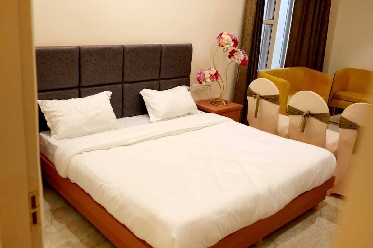 A neatly made bed with white linens, a dark headboard, and a small bedside table with a vase of flowers. There are two chairs with yellow upholstery and white headrests.