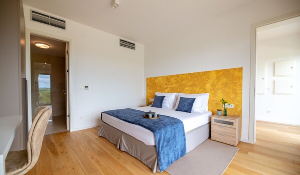 Ein modernes Schlafzimmer mit einem großen Bett, einem Nachttisch, einem Sessel und Blick ins Freie.
