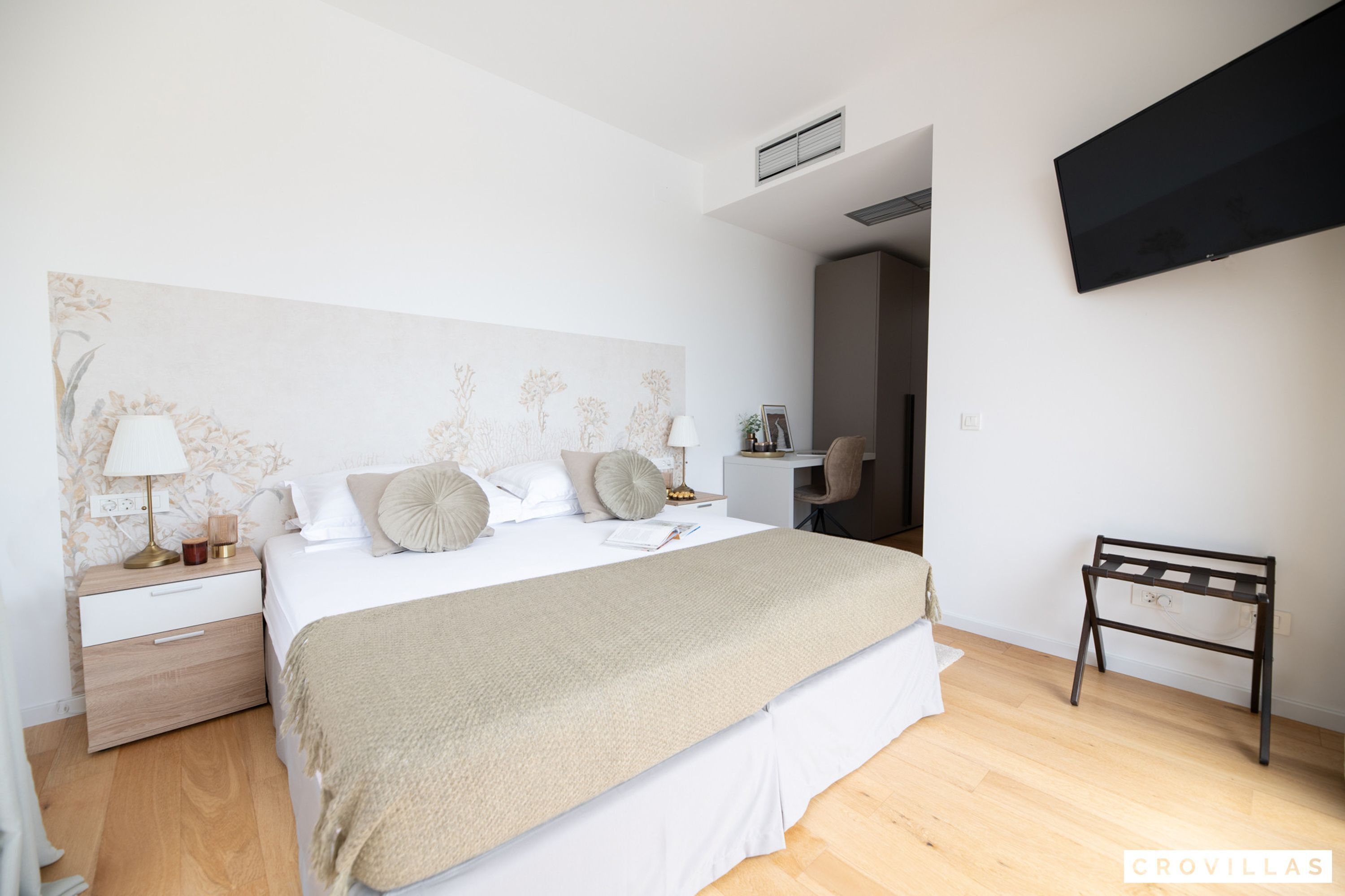 Un dormitorio con cama, mesita de noche, lámpara y televisor montado en la pared.