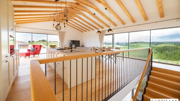 Une cuisine spacieuse avec un grand îlot central, des poutres en bois au plafond et une vue sur la campagne grâce à de grandes fenêtres.