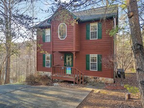 A two-story wooden house with green shutters, a stone foundation, and a front porch with a bench.