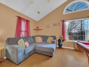 Una sala de estar con un sofá seccional gris, piso de madera, una ventana con vista a árboles y un ventilador de techo.