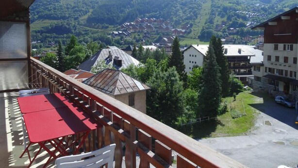 A balcony with a red table and chairs, overlooking a village with buildings and a mountain in the background. - Resort center, 3rd floor, balcony, parking, 53m², Valloire (Valloire)