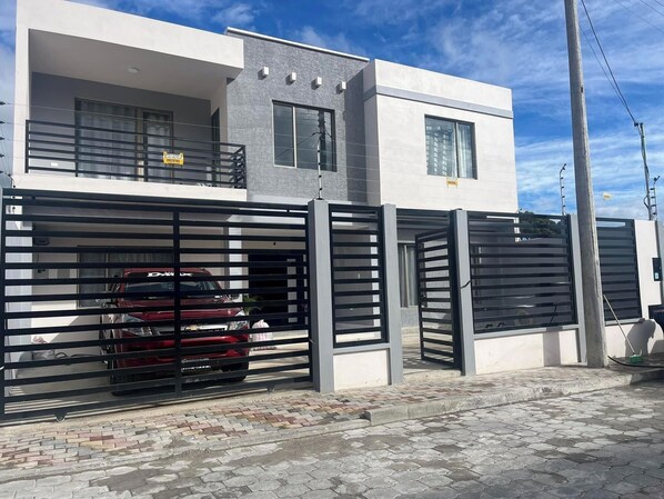 Un edificio moderno de dos pisos con entrada protegida por una reja, un auto rojo estacionado adentro y un camino de entrada pavimentado.