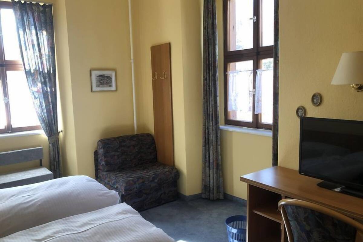 Ein Hotelzimmer mit zwei Betten, einem Schreibtisch mit Fernseher, einem Stuhl, einem Fenster mit Vorhängen, einer Tür und einem Bild an der Wand.