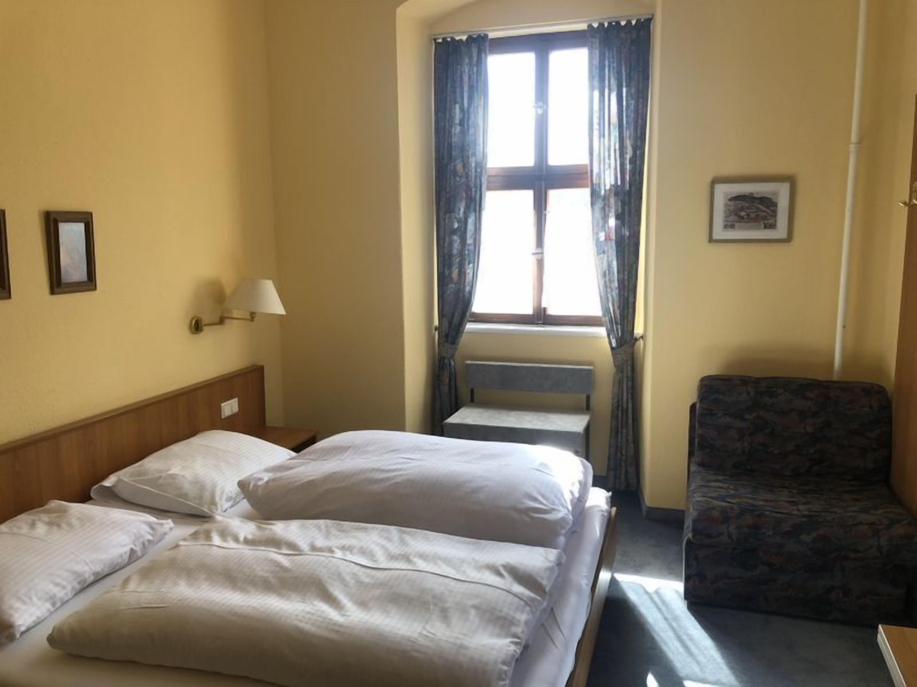 Une chambre d’hôtel avec un lit, une chaise, une fenêtre avec des rideaux, une lampe et un tableau encadré au mur.