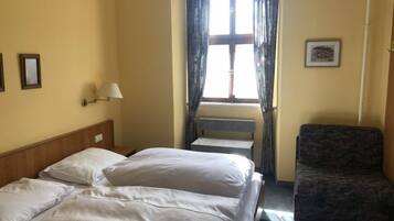 Ein Hotelzimmer mit einem Bett, einem Sessel, einem Fenster mit Vorhängen, einer Lampe und einem gerahmten Bild an der Wand.