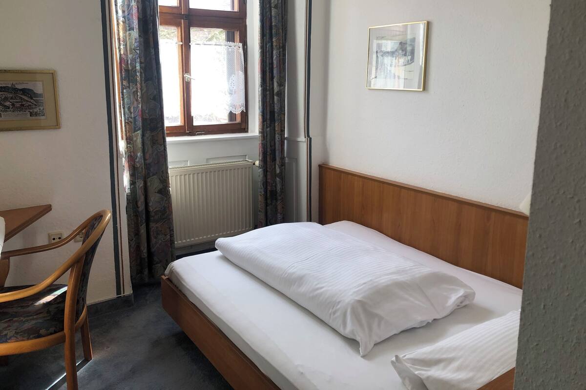 Ein kleines, ordentlich eingerichtetes Schlafzimmer mit einem Einzelbett, einem Sessel, einer Heizung und einem Fenster mit Vorhängen.