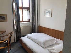 A small, neatly arranged bedroom with a single bed, a chair, a radiator, and a window with curtains. - Gasthaus Klosterhof (Wertheim)