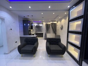 Un salon moderne avec deux fauteuils noirs, une cuisine en arrière-plan et un meuble mural décoratif avec des étagères.