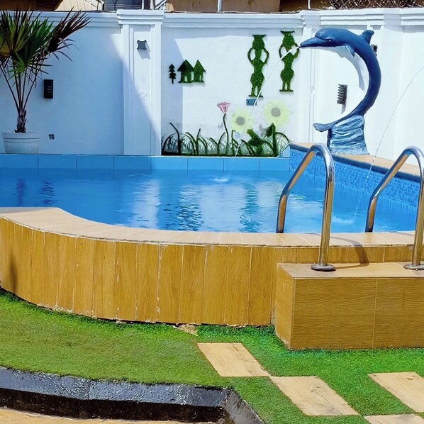 A swimming pool with a dolphin sculpture, surrounded by a wooden deck and artificial grass. - Gilllant Luxury Homes Apartment (Benin City)