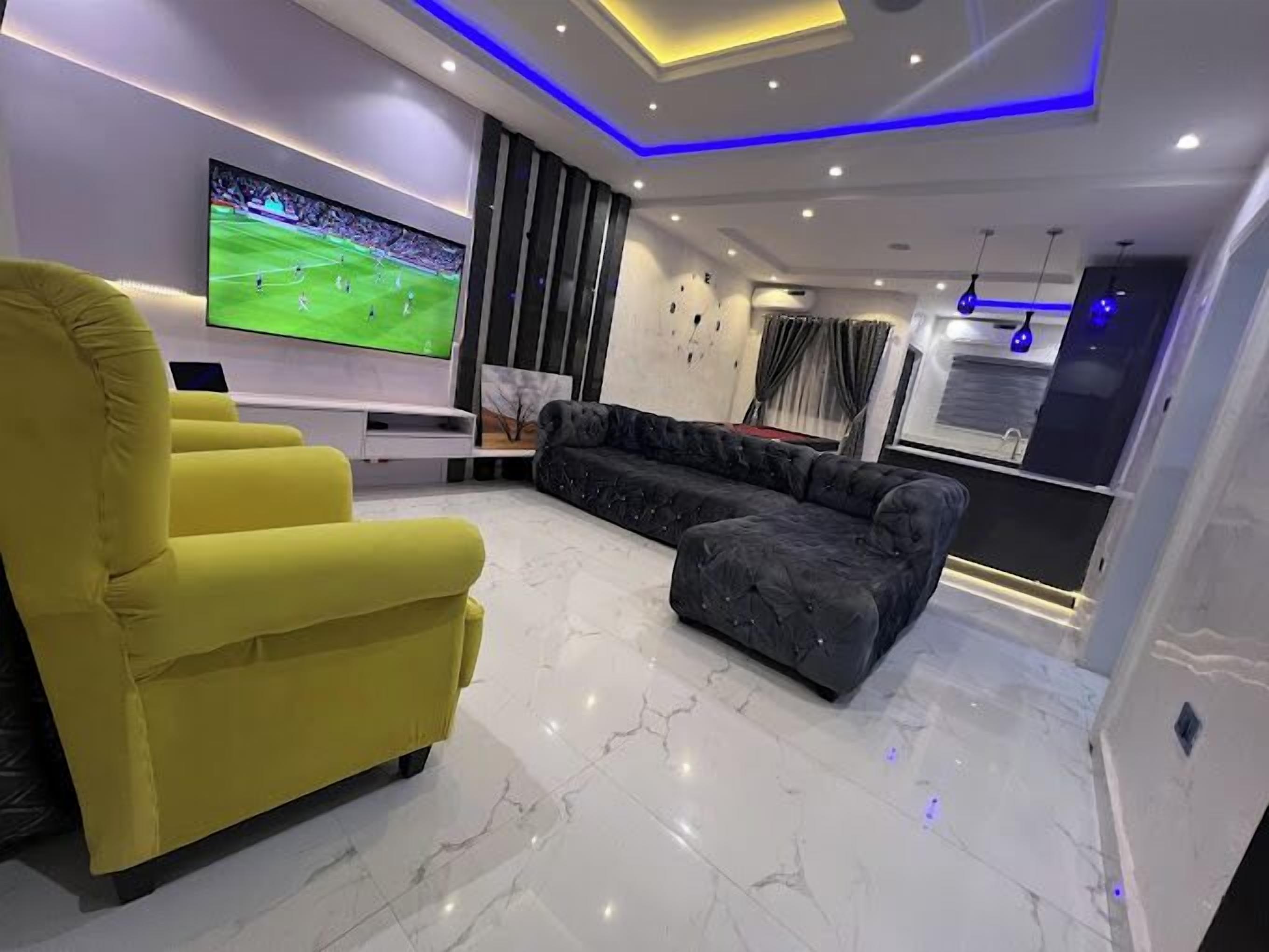 Una sala de estar moderna con un televisor de pantalla plana grande, un sillón amarillo, un sofá oscuro y piso de mármol.