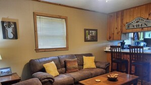 Ein gemütliches Cabin-Interieur mit einem braunen Ledersofa, einem Holztisch und einer Küchenzeile mit Holzschränken und einem Schild mit der Aufschrift 'Welcome to Cabin'.