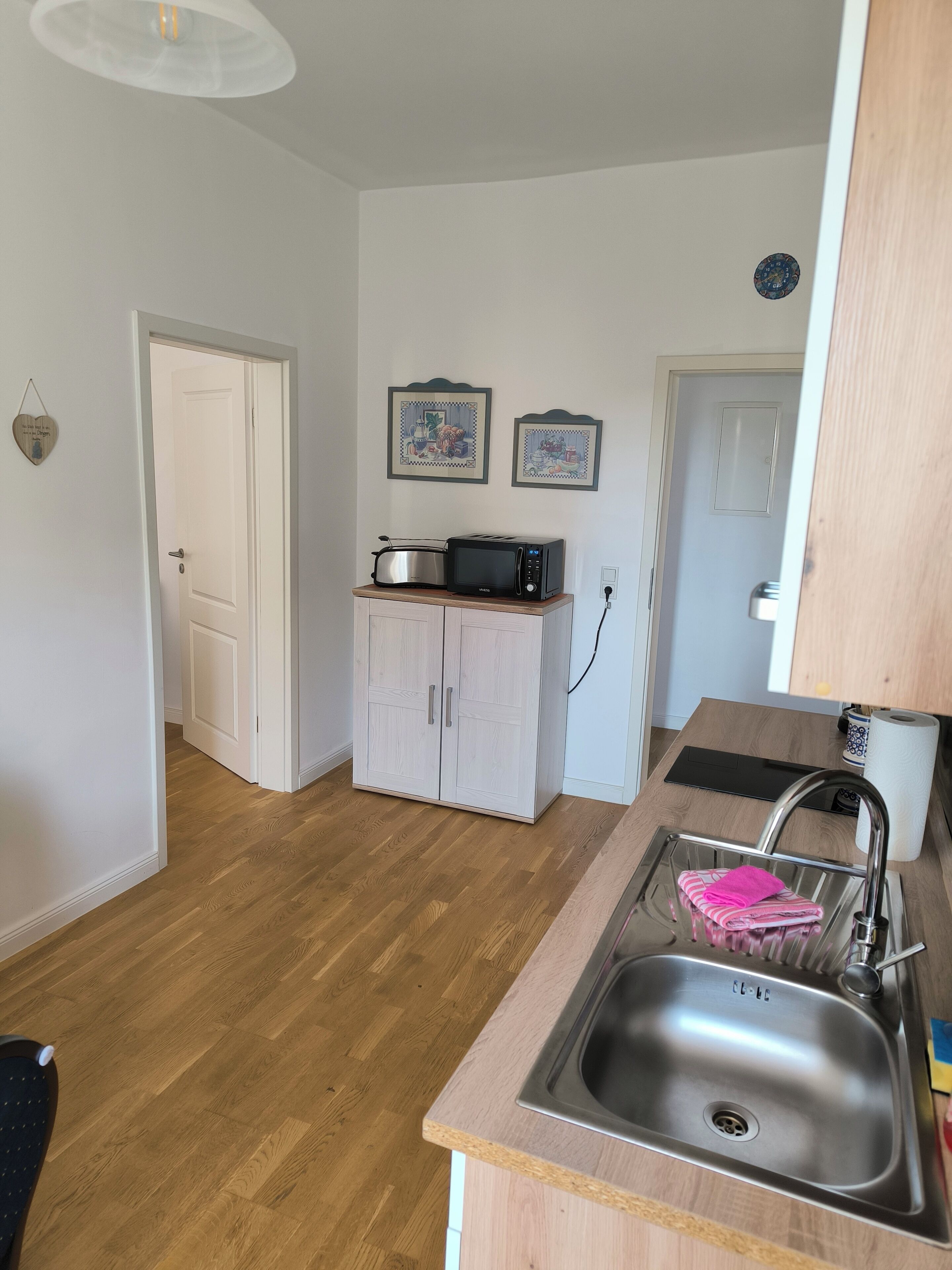 Une cuisine de taille réduite, dotée d’un plan de travail en bois, d’un évier en acier inoxydable et d’un four à micro-ondes. Deux tableaux encadrés ornent le mur, et une porte donne accès à une autre pièce.