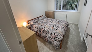 Une chambre à coucher comprenant un lit, une table de chevet avec une lampe, une commode, une fenêtre avec des stores et un radiateur.