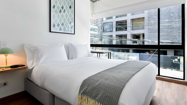 Une chambre d’hôtel moderne avec un grand lit, une table de chevet avec une lampe, une œuvre d’art encadrée au mur et une vue sur la ville grâce à de grandes fenêtres.