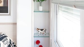 A room with a white wall, a framed artwork, a glass shelf with decorative items, and a window with blinds. - Unik Vacation Zamenhoff (Tarragona)