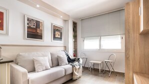 A modern living room with a beige sofa, a small table, and a folding chair. There are framed artworks on the wall and a window with blinds. - Unik Vacation Zamenhoff (Tarragona)