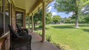 A covered porch with wicker chairs, a brick building, and a grassy lawn with trees. - Southall Cottage: 3bd*Serene Outdoor Space* (Franklin)