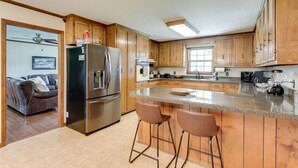 A kitchen with wooden cabinets, a stainless steel refrigerator, a large island with bar stools, and a dining area with a sofa and a ceiling fan. - Southall Cottage: 3bd*Serene Outdoor Space* (Franklin)