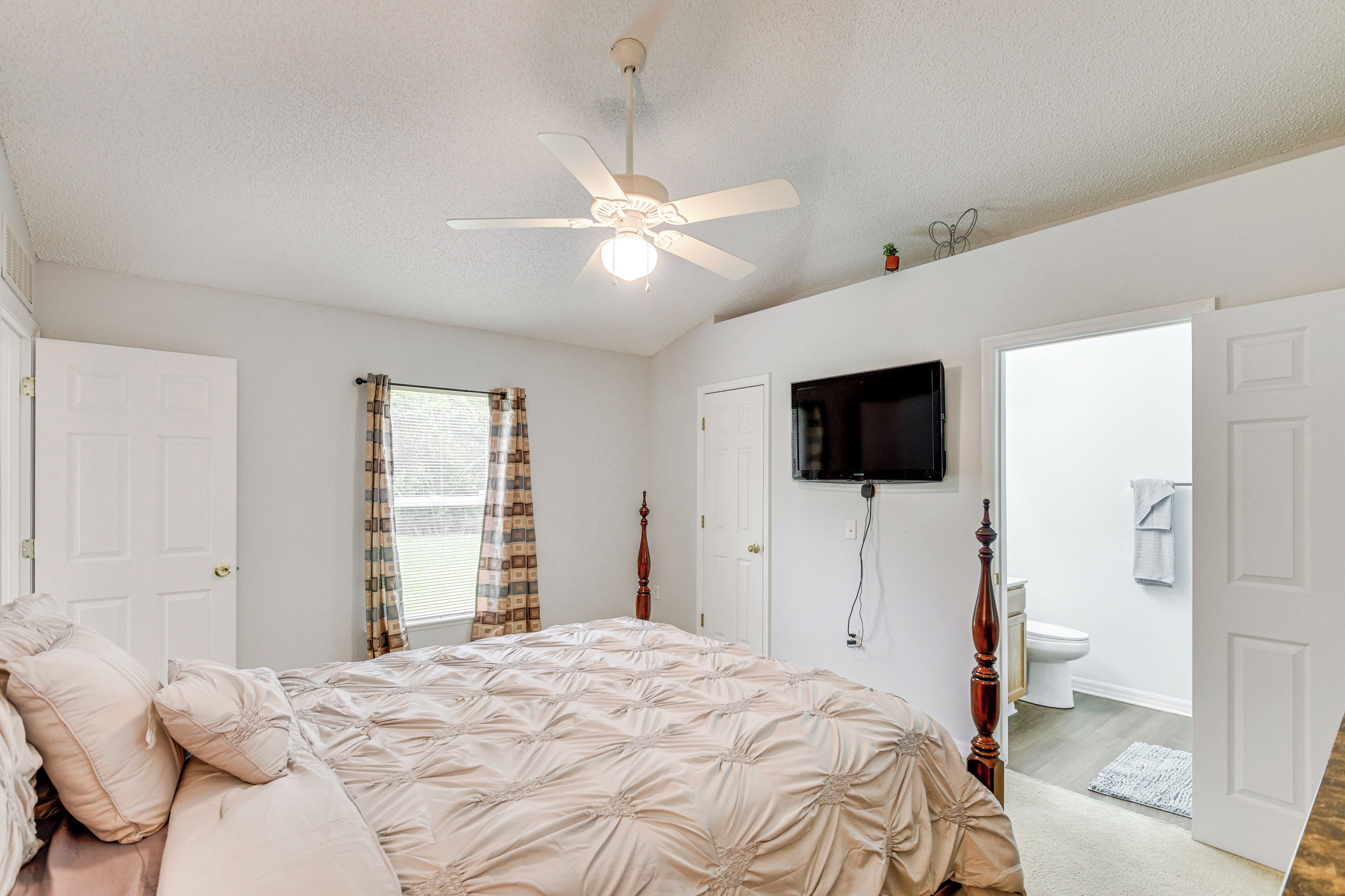 Ein Schlafzimmer mit Bett, Deckenventilator, an der Wand montiertem Fernseher und einem durchgegangene Tür sichtbaren Badezimmer.