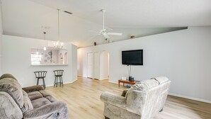 Amplia sala de estar con un sofá gris, un sillón blanco y una mesa de centro de madera. Un ventilador de techo y un televisor de pantalla plana están montados en la pared.