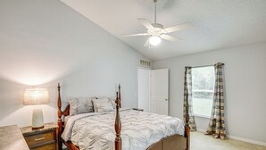 Un dormitorio con cama con dosel, una mesita de noche con lámpara, ventilador de techo, ventana con cortinas y una puerta.