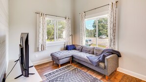 A living room with a sectional sofa, a flat-screen TV, and large windows with curtains. - 3 Mi to Duke: Downtown Durham Cottage! (Durham)