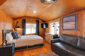 A cozy wooden cabin bedroom with a bed, a black leather sofa, a TV, and a painting on the wall. - Pet-friendly Apple Valley Getaway w/ Fire Pit! (Hurricane)