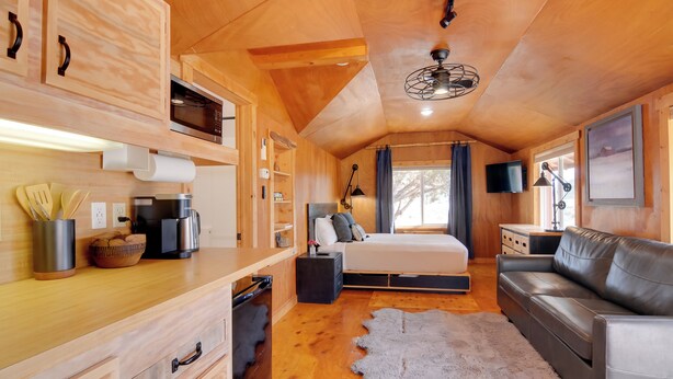 Un accogliente interno di una baita con soffitto in legno, un letto con cuscini, una poltrona in pelle, un'area pranzo con tavolo e sedie, e un'ampia finestra.