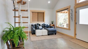Un salon moderne avec un canapé d’angle gris, une grande fenêtre offrant une belle vue, une échelle en bois et une fougère en pot.