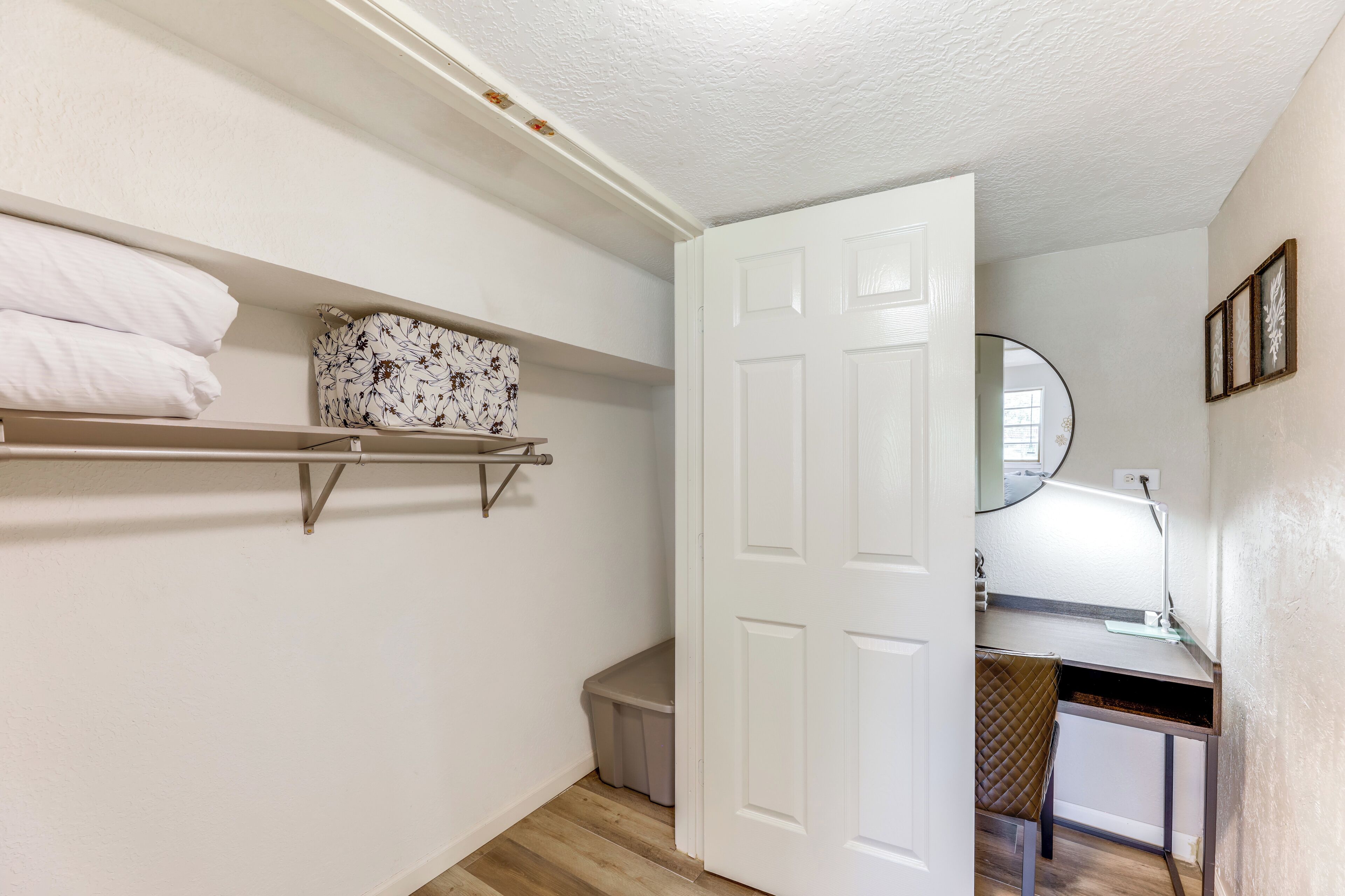 A small, clean room with a bed, a bedside table, a nightstand, and a wall-mounted shelf with pillows and a decorative item.
