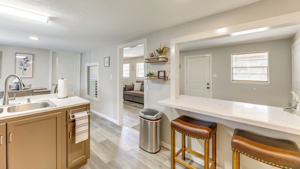 A modern kitchen with a sink, countertop, and stools. There is a trash can and a window with blinds. - Somerville Lake House - Walk to Water! (Somerville)