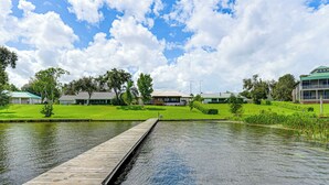 A wooden dock extending into a calm body of water with a grassy area and buildings in the background. - Waterfront Home w/ Boat Dock in Lake Placid! (Lake Placid)