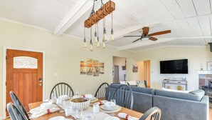 Une salle à manger avec une table en bois dressée pour six personnes, entourée de chaises. On aperçoit un ventilateur de plafond et un téléviseur à l’arrière-plan.