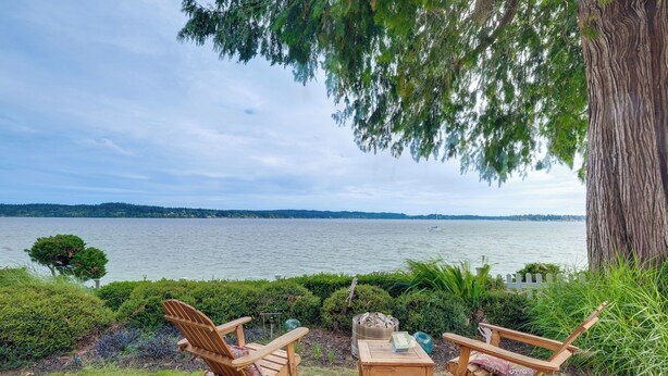 A lakeside view with two wooden chairs and a table, surrounded by greenery and a large tree. - Studio w/ Bay View: Beach Access! Octopus's Garden (Bremerton)