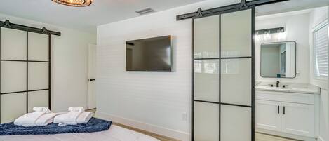 A modern bedroom with a bed, a sliding door closet, a mounted TV, and a bathroom with a sink and mirror.