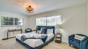 Une chambre avec un grand lit, deux fenêtres équipées de stores, une table de chevet avec une lampe, une chaise bleue et un luminaire fixé au mur.