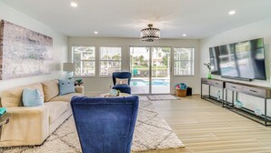 Un salon comprenant un canapé, un fauteuil bleu, un téléviseur et une grande fenêtre donnant sur une piscine.
