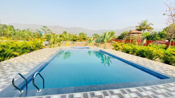 A rectangular swimming pool with a tiled deck, surrounded by lush greenery and palm trees, with mountains in the background. - Shanti Forest Resort (Karjat)