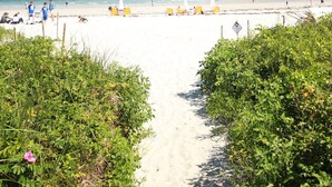 En sti af sand der fører ned til havet, med strandstole og parasoller.