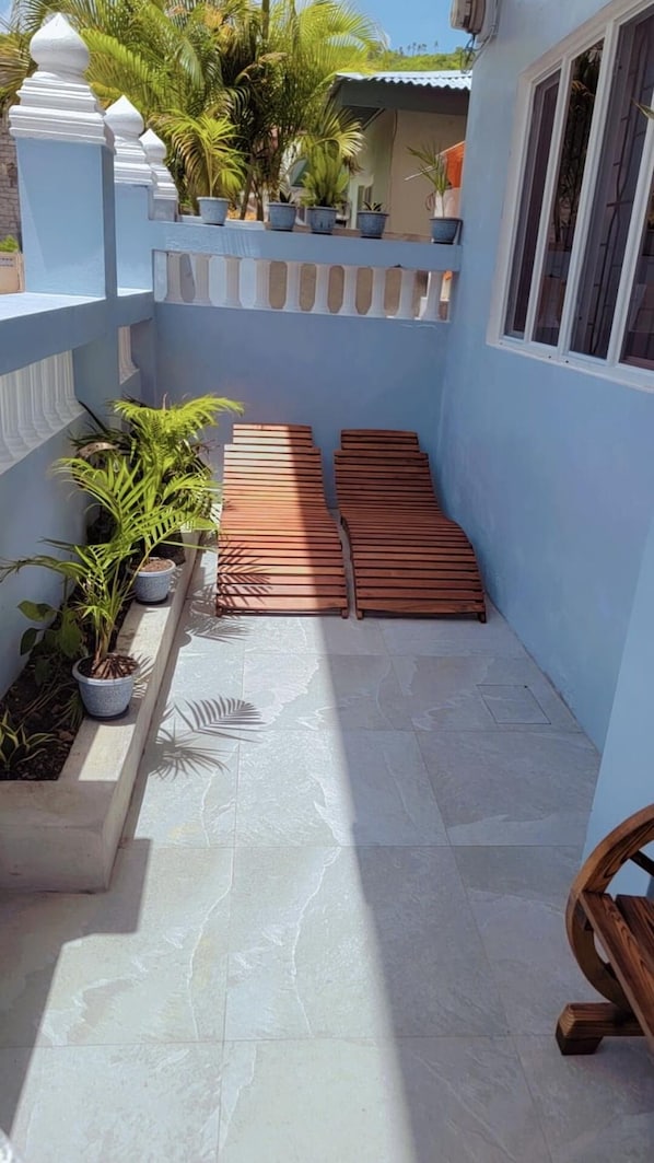 A narrow outdoor area with a tiled floor, wooden benches, potted plants, and a white railing.