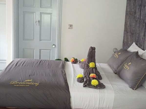 A hotel room with a bed covered in a grey blanket, pillows with 'Dreaming' embroidered, and a towel with flowers.