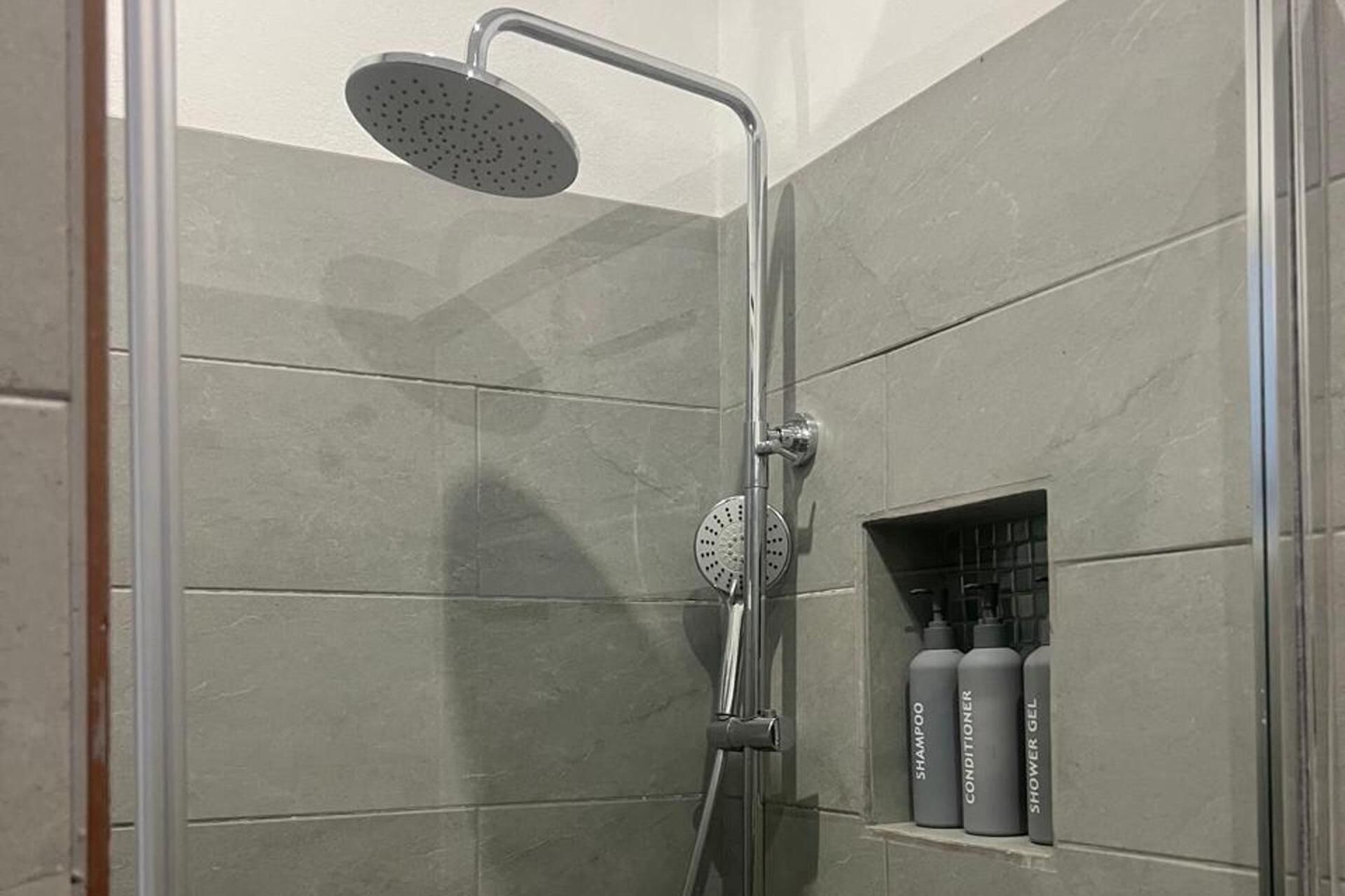 En bruser med håndbruser og betjening på væggen. Tre flasker shampoo, balsam og shower gel står på en hylde.