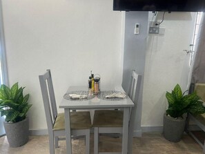 A dining area with a table set for two, two chairs, a TV mounted on the wall, and potted plants on either side. - Lovely Studio Apartment in Soufriere From Claviers (Soufrière)