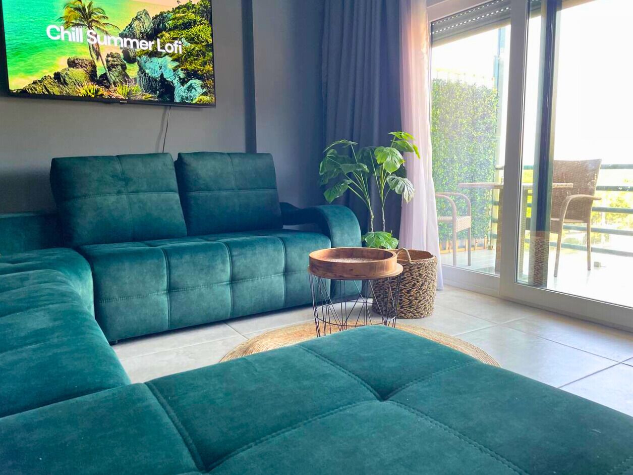 A modern living room with a teal sectional sofa, a small wooden side table, and a large window offering a view of the outdoors.