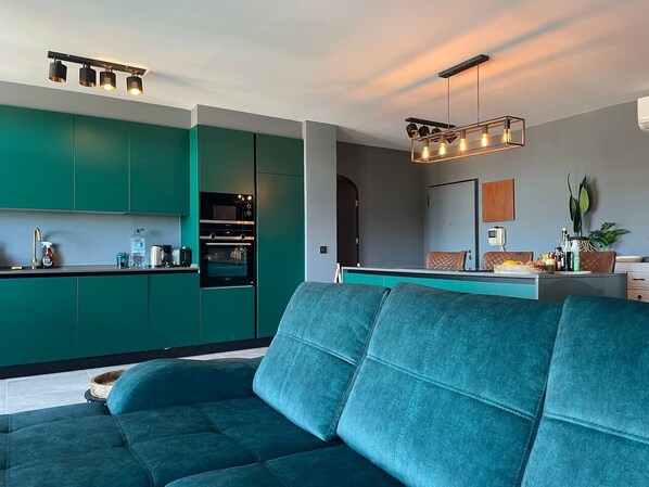 A modern kitchen with teal cabinetry, a built-in oven, and a dining area with a teal sofa. - Caparica Surf Retreat (Almada)