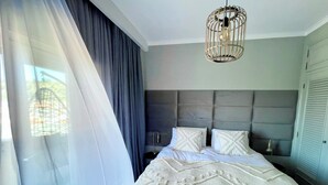 A bedroom with a bed, a hanging light fixture, a nightstand, and a window with sheer curtains. - Caparica Surf Retreat (Almada)